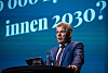 Bjørnar Skjæran står ved talerstol med vannkaraffel foran blå konferansebakgrunn.